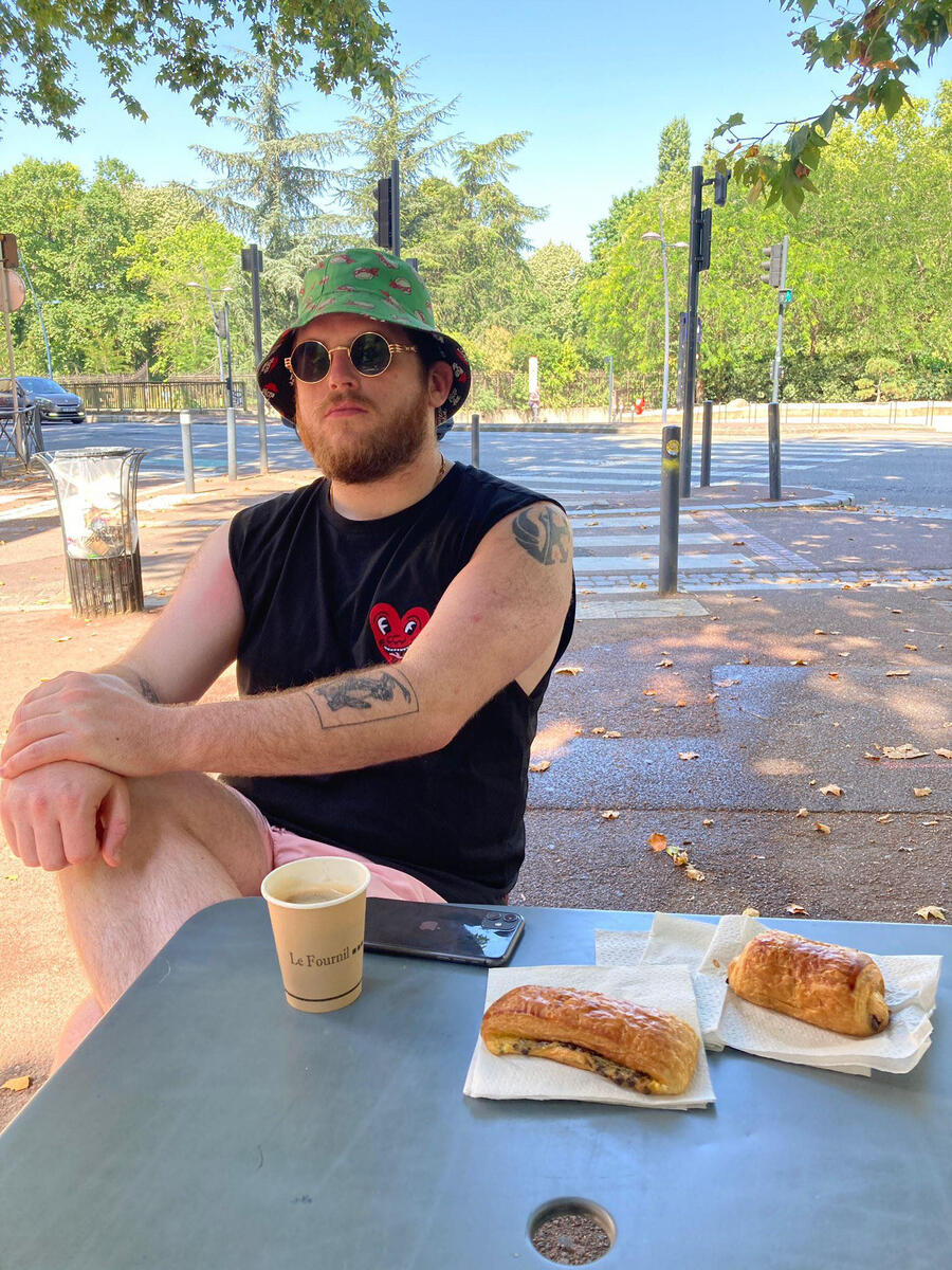 My official author pic. I am sitting with my legs crossed wearing a sleeveless Keith Haring top, pink swimming shorts and a TMNT bucket hat. A coffee, my phone and two pastries are on the table in front of me. Pic inspired by an Anthony Bourdain picture.